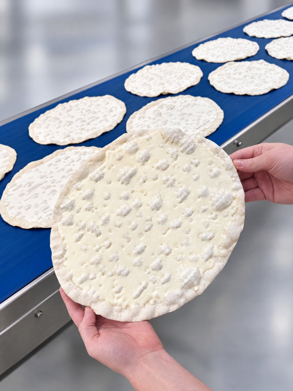 Pizza bases with a thin layer of white sauce on a blue belt, one base held up in two hands