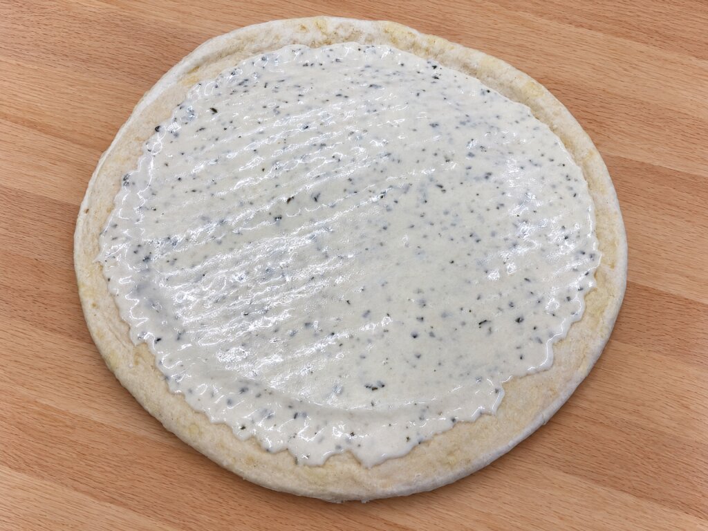 Round pizza base with a layer of white herb sauce, seen from above on a wooden board
