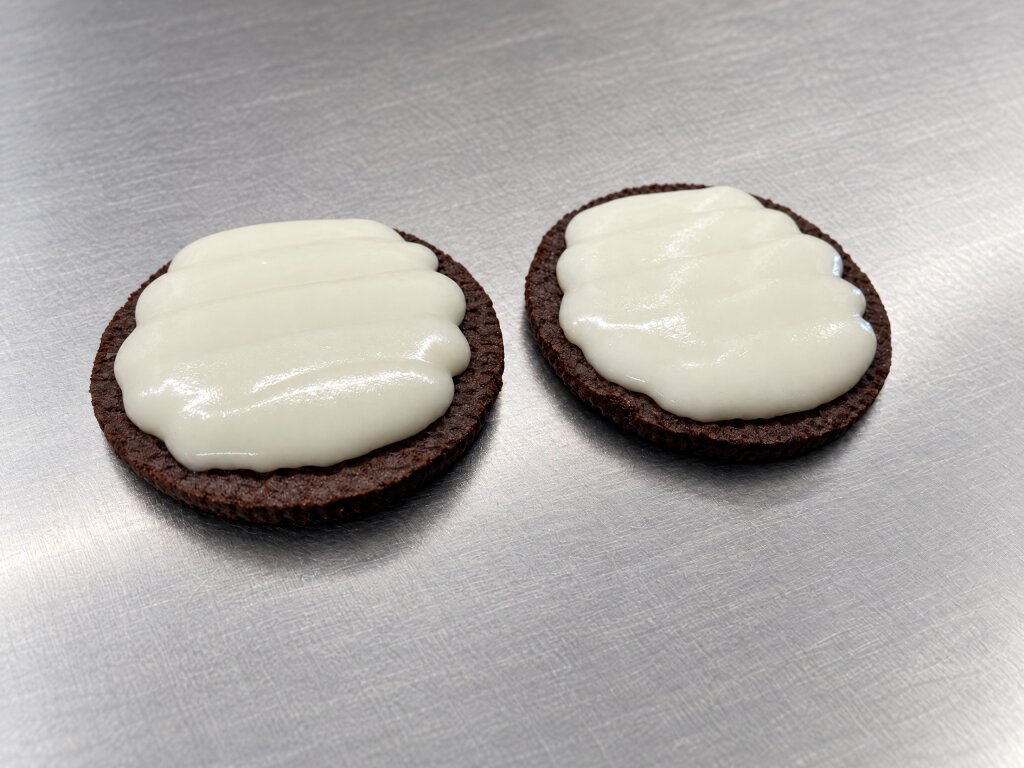 Two cocoa biscuits with a layer of white cream deposited on top, on stainless steel