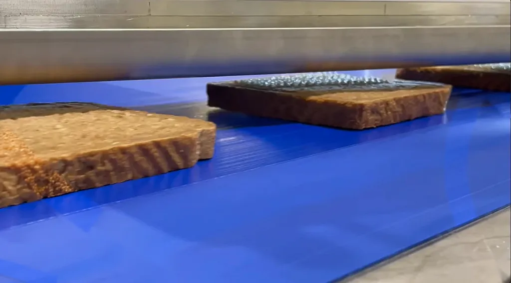 Slices of brown bread on a blue conveyor under the machine, the middle slice with rows of small butter drops on top