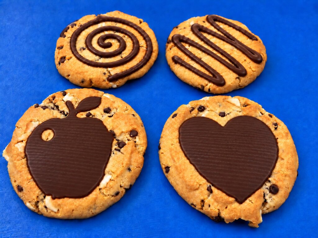 Four cookies with dark chocolate shapes on a blue background: spiral, zigzag line, apple and heart